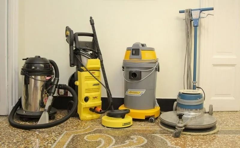 Attrezzatura per la pulizia: aspirapolvere, idropulitrice, lavasciuga e lucidatrice per pavimenti, in ambienti chiusi, su un pavimento decorato contro una parete bianca.