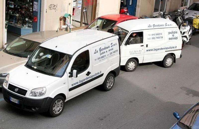 Due furgoni bianchi per le consegne parcheggiati in una strada cittadina, con la scritta