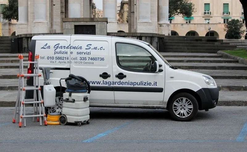 Furgone bianco per le pulizie parcheggiato di fronte a una struttura in pietra, con scala e aspirapolvere.