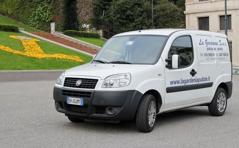 Furgone Fiat Doblò bianco con scritta sulla fiancata, parcheggiato su un'area asfaltata con un pendio erboso e un edificio sullo sfondo.