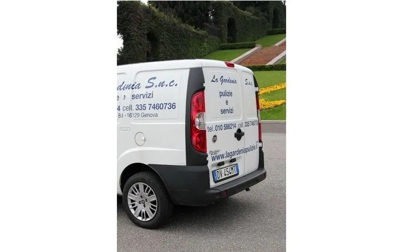 Furgone bianco con loghi e scritte parcheggiato vicino a un giardino con sentiero e vegetazione.