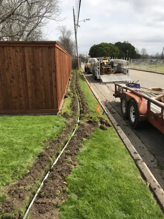 Roadside construction beside a wooden fence, with a trench in the grass and workers near heavy equipment.