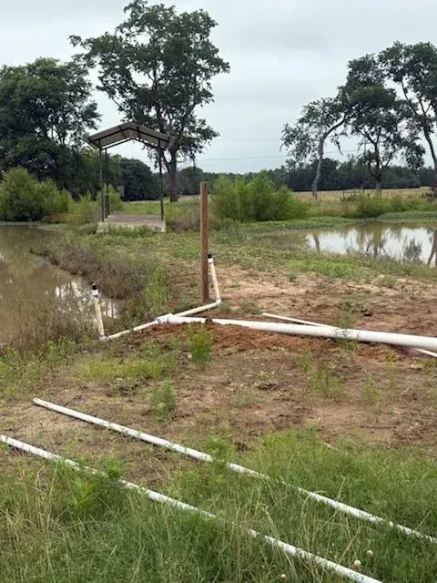 Rural pond beside grassy field with a small shelter and scattered white PVC pipes in the foreground