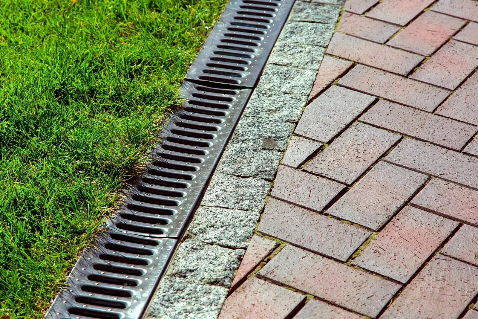Drain grate between green grass and a brick-paved walkway