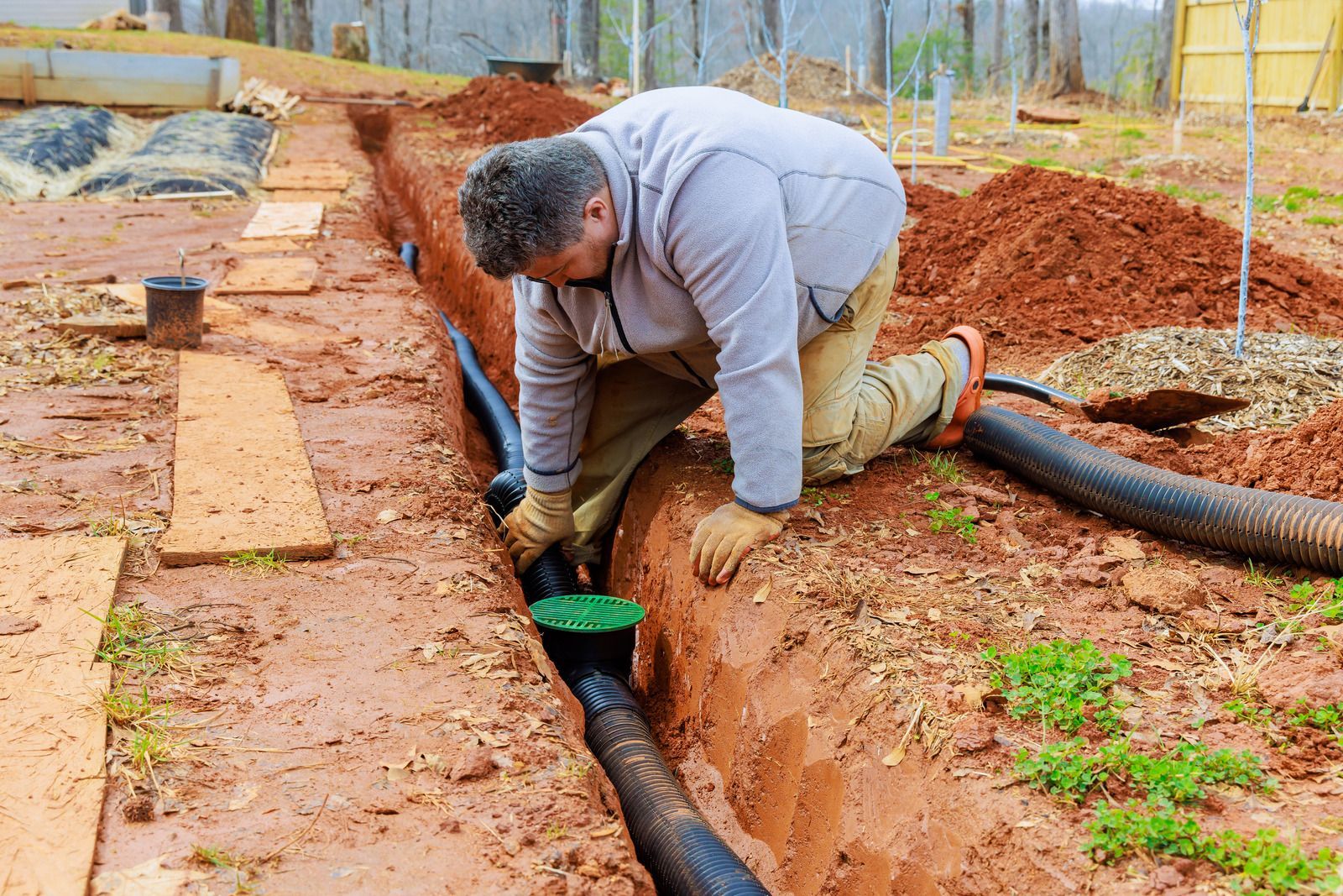 Drainage trench lined with white fabric, filled with gravel and a black corrugated pipe beside a garden bed