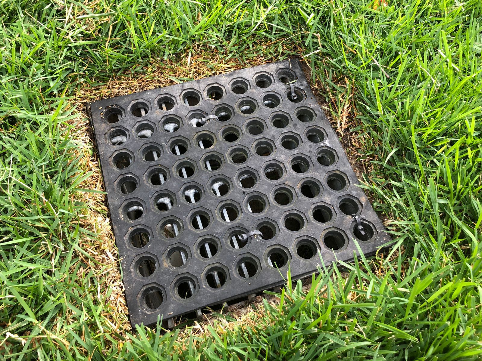Green-topped outdoor drain box with black pipe and white corrugated pipe in a gravel bed on grass