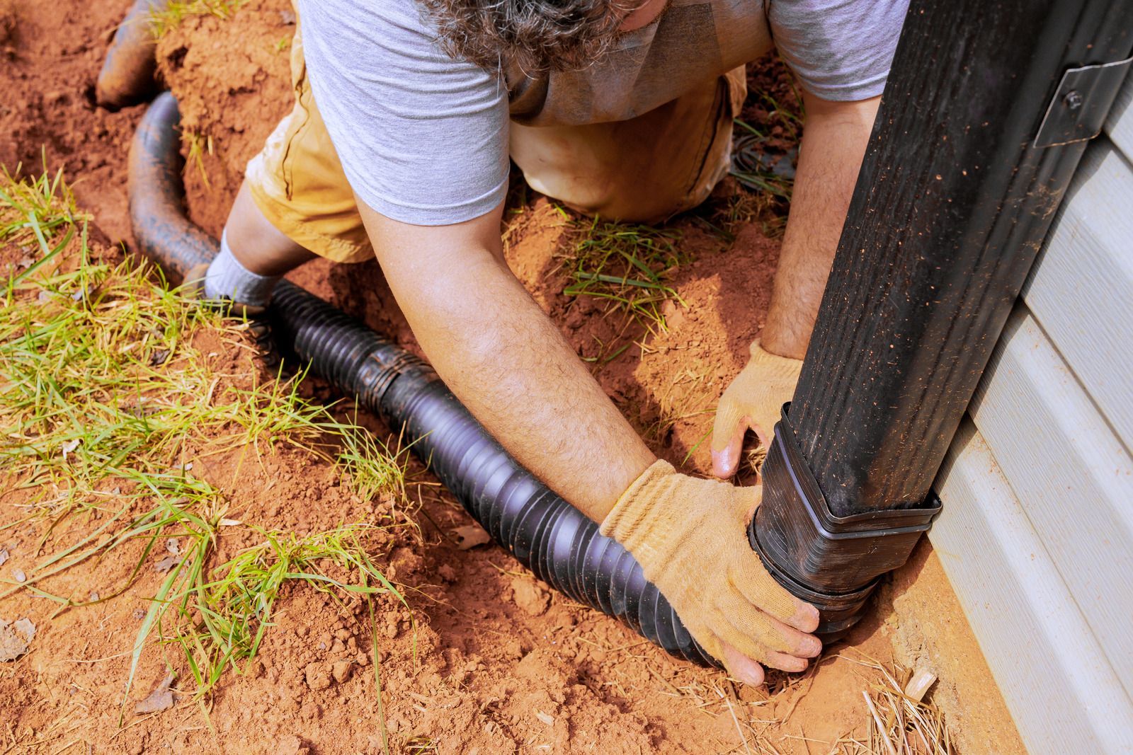 Person connecting black drainage pipe to a downspout in a dirt yard.