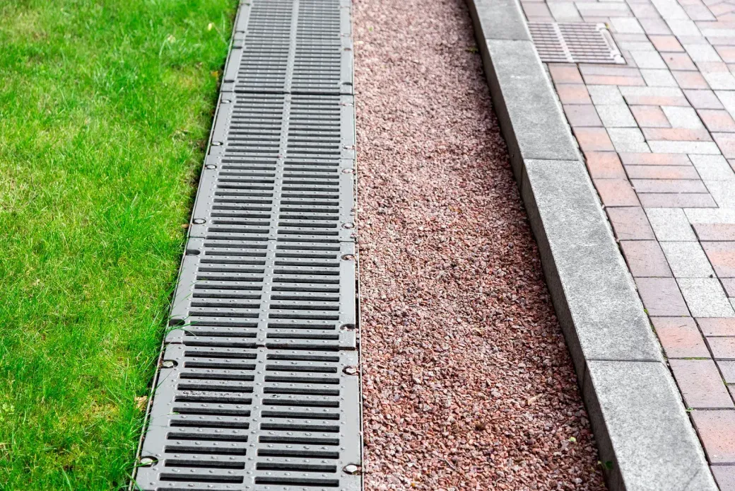 Metal drainage grate beside grass, red gravel, and a paved walkway