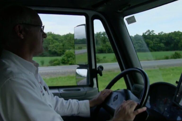 Driver at the wheel of a truck, looking out over a green roadside landscape.