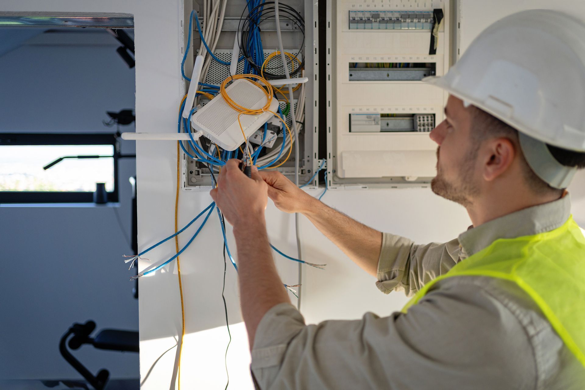 Un elettricista sta cablando un quadro elettrico aperto, con cavi blu e arancioni, in una luminosa stanza industriale.