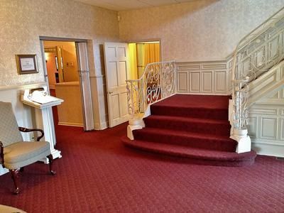 Red-carpeted entrance hall with ornate staircase, open doorways, and a white pedestal with a book and chair.
