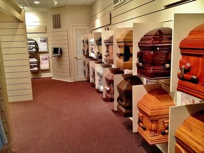 Row of closed caskets on display in a funeral home showroom. Various wood finishes and styles. Brown carpet.