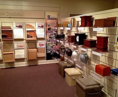 Display of cremation urns and boxes in a funeral home. Various sizes, colors, and materials. Wooden shelves on the wall.