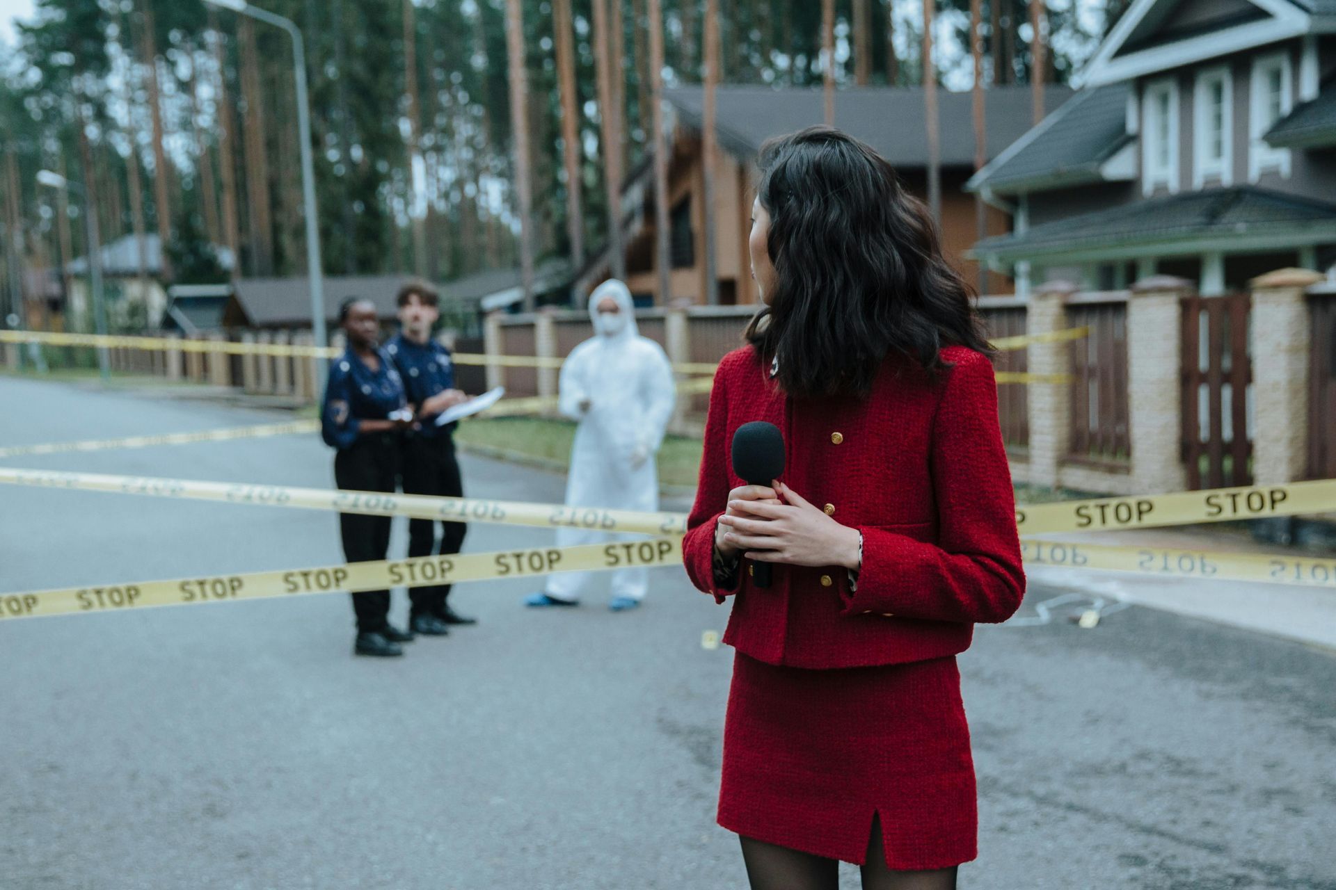 A reporter in a red suit holds a microphone in front of a street cordoned off by police and a person in a hazmat suit.