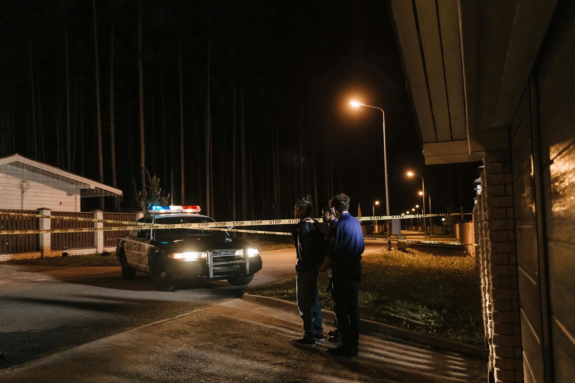 Two people stand near a police car at night, cordoned off by yellow police tape in a residential area.