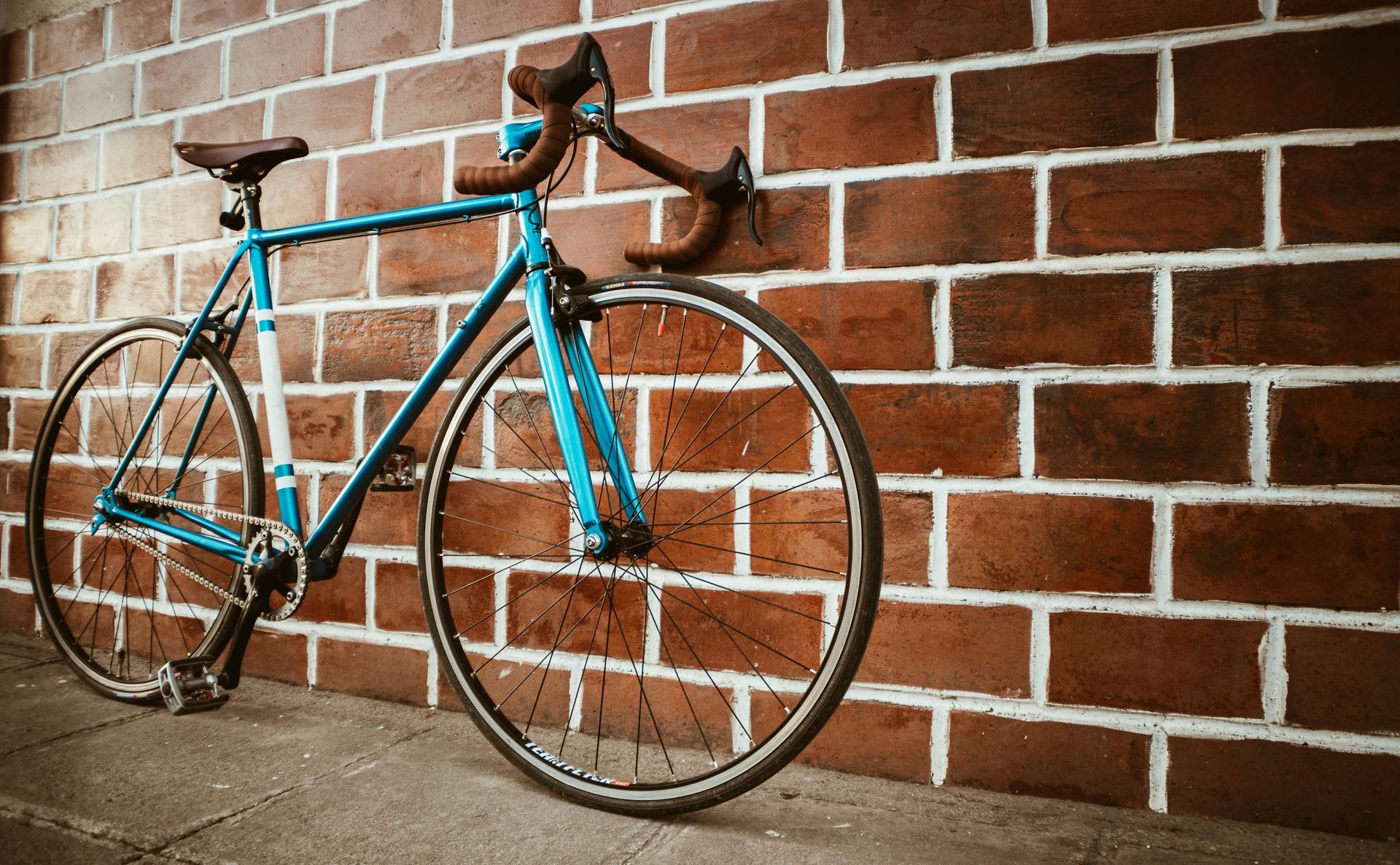 A turquoise vintage road bike with brown handlebars stands against a red brick wall.