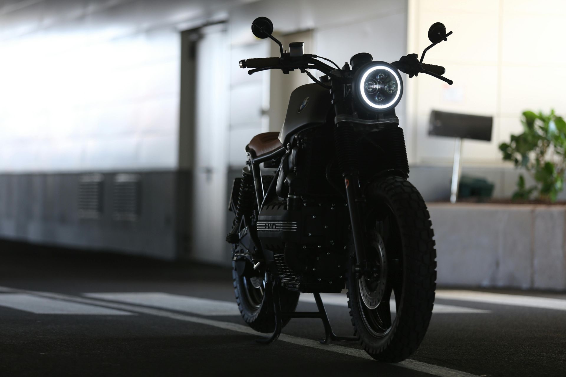 A black cafe racer-style motorcycle parked in an indoor garage with its circular LED headlight illuminated.