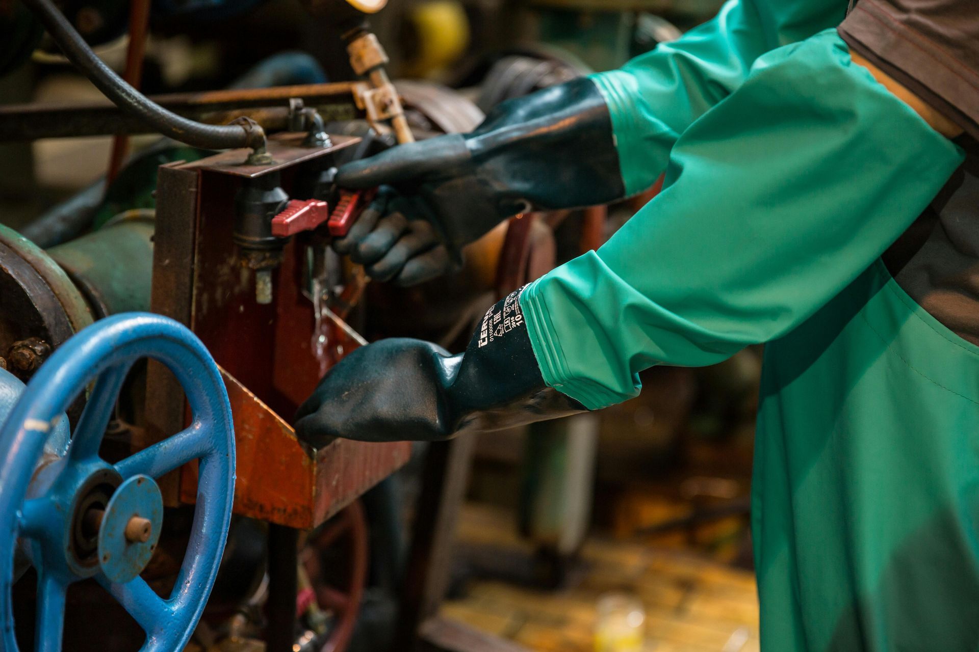 A worker in a green protective apron and black rubber gloves operates industrial machinery with manual levers.