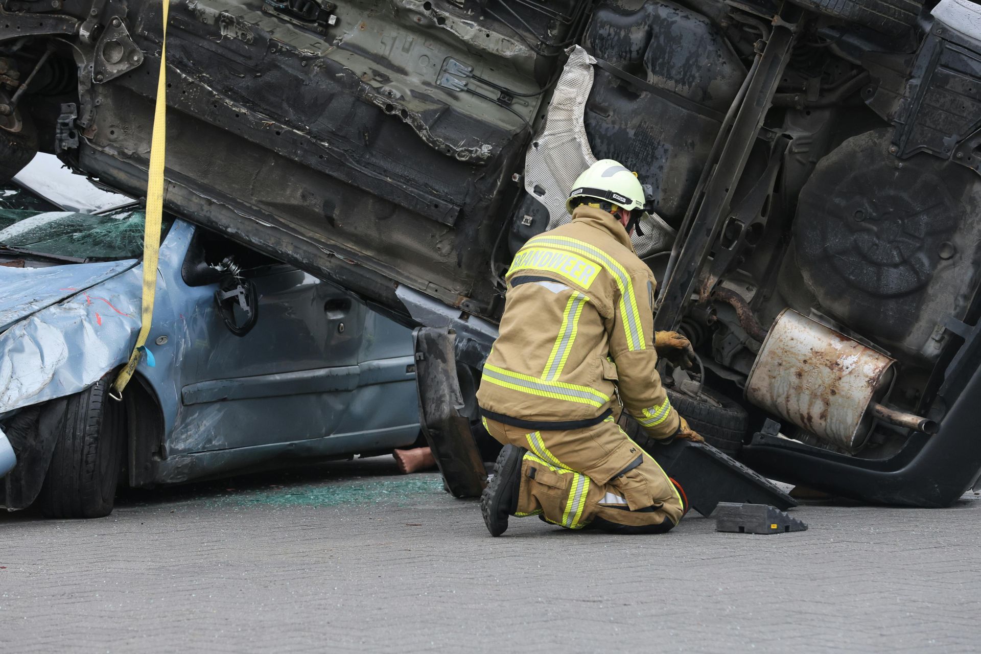 A firefighter in tan protective gear kneels on pavement near a wrecked car partially underneath another overturned vehicle.