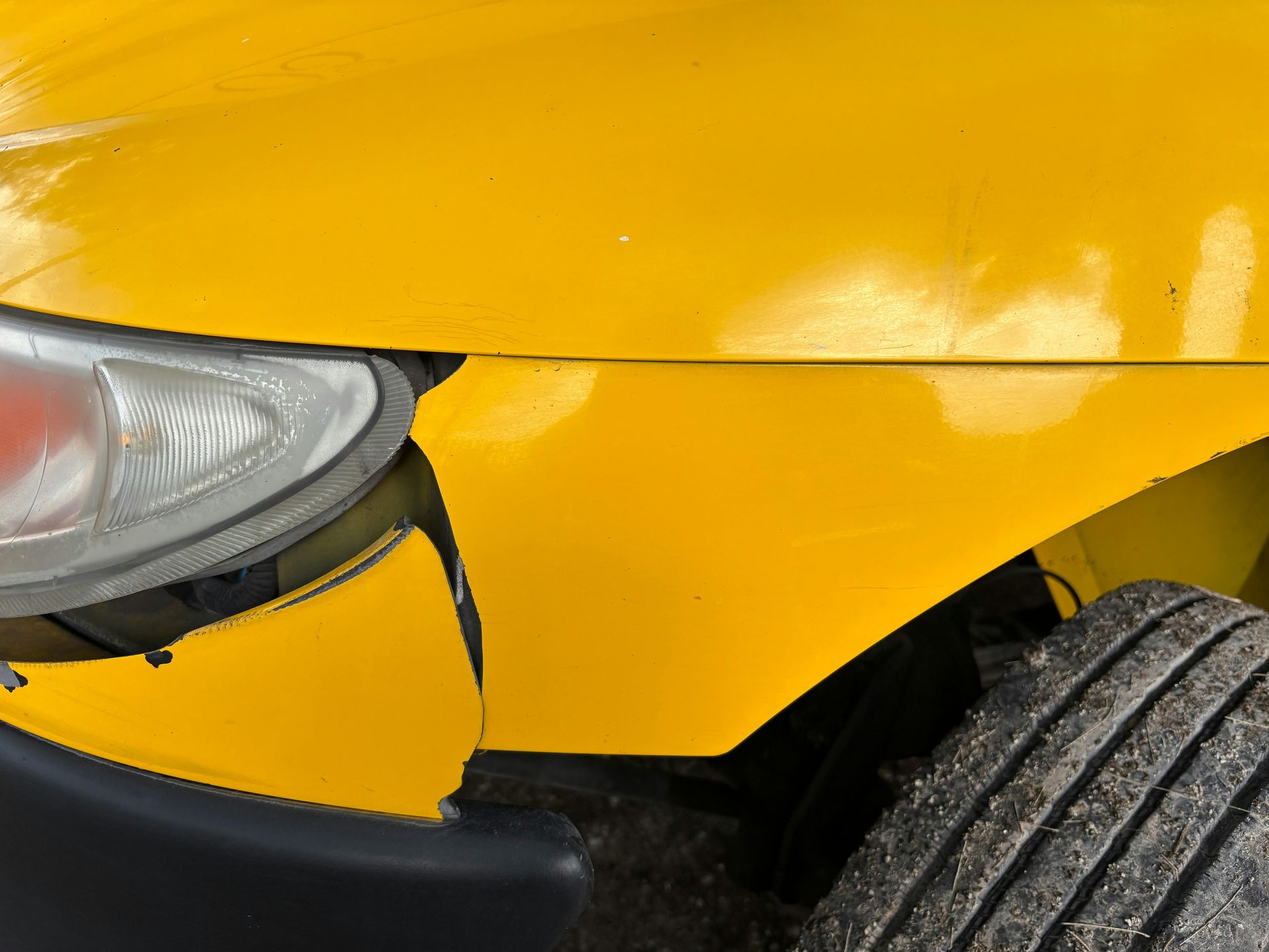 A close-up view of a yellow vehicle with a misaligned, damaged front bumper separated from the headlight and fender.