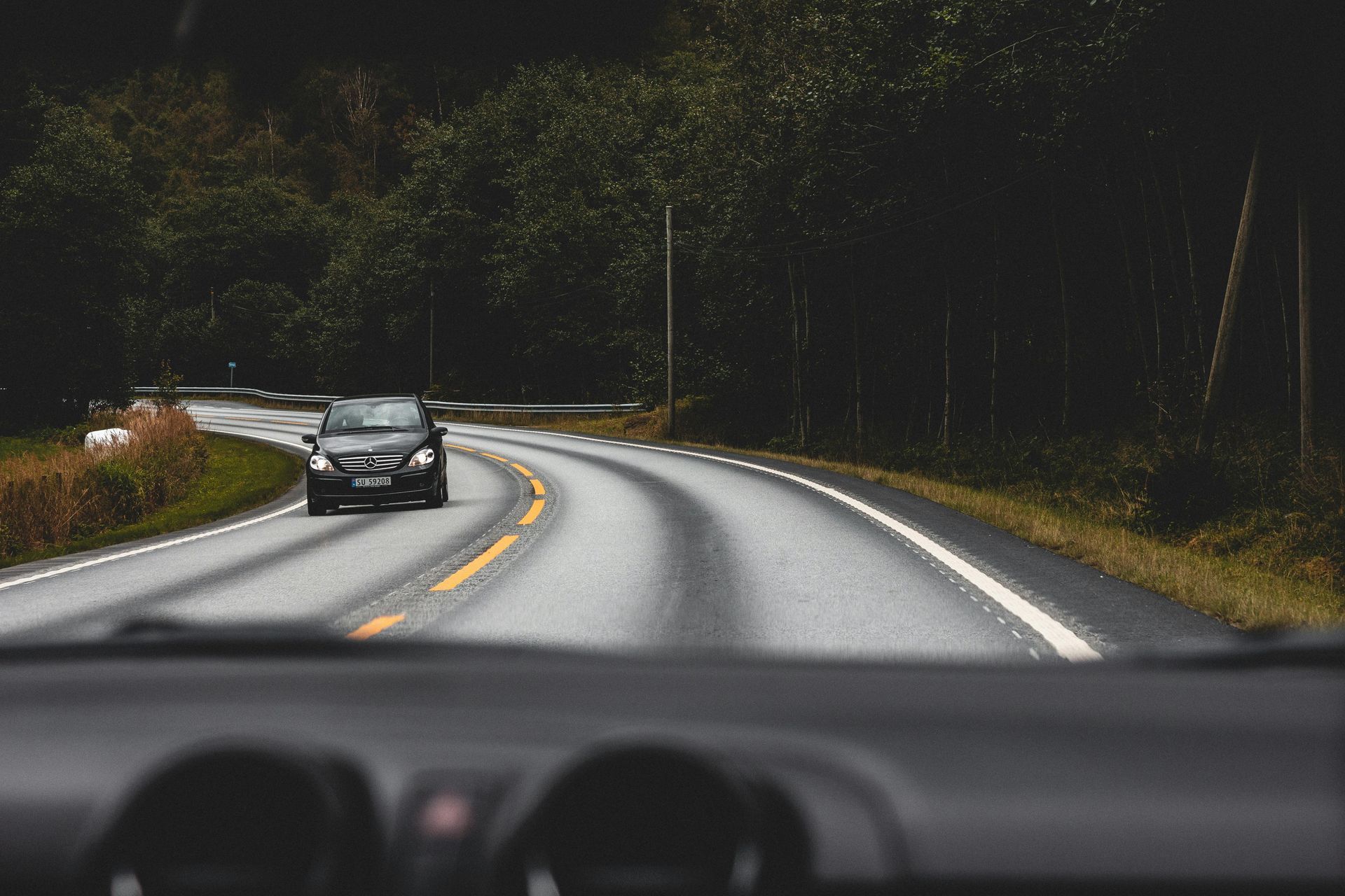 A black car drives toward the viewer on a gray asphalt road lined with trees.