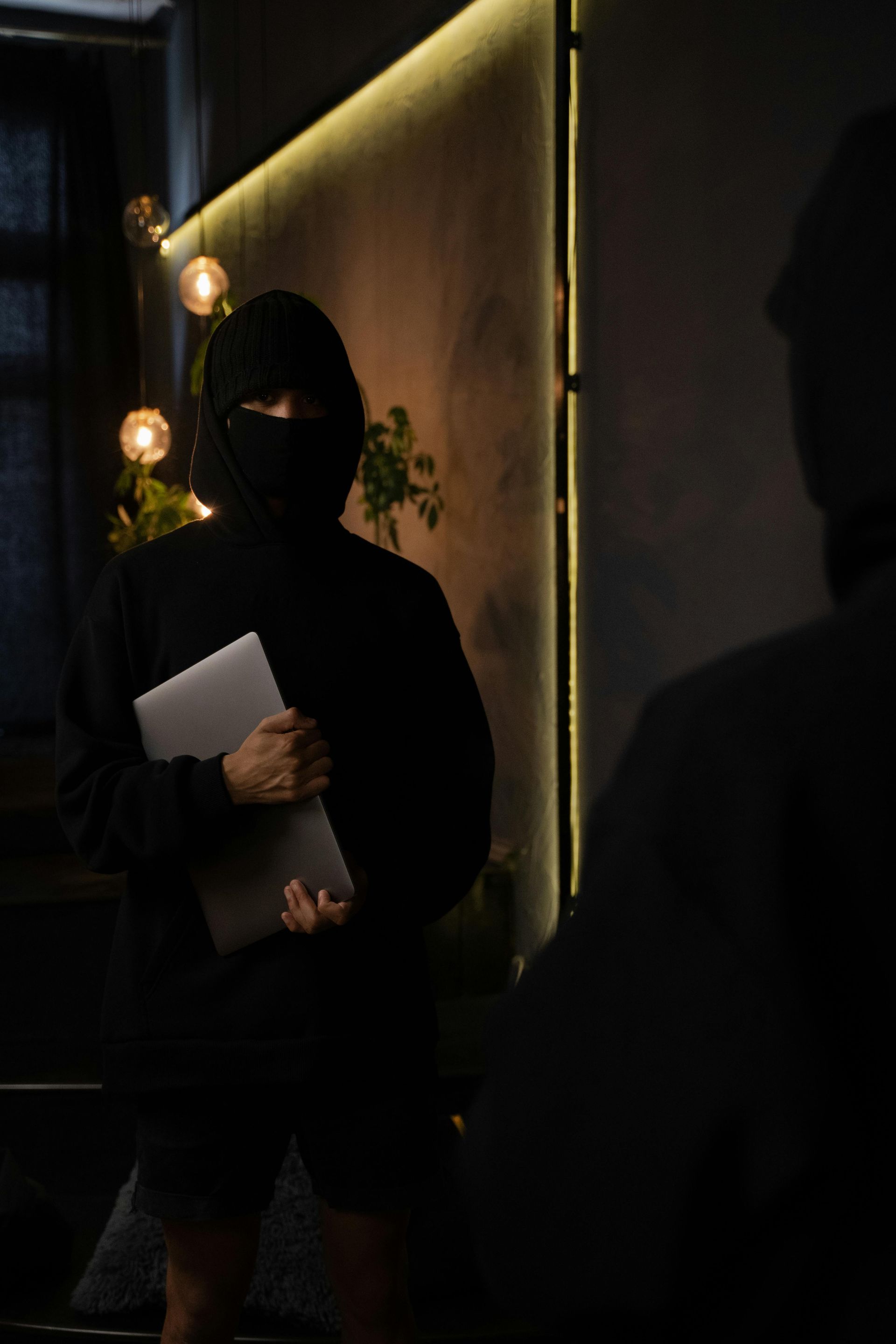 A person in a black hoodie and face mask holding a closed laptop, standing in a dimly lit room with decorative lighting.