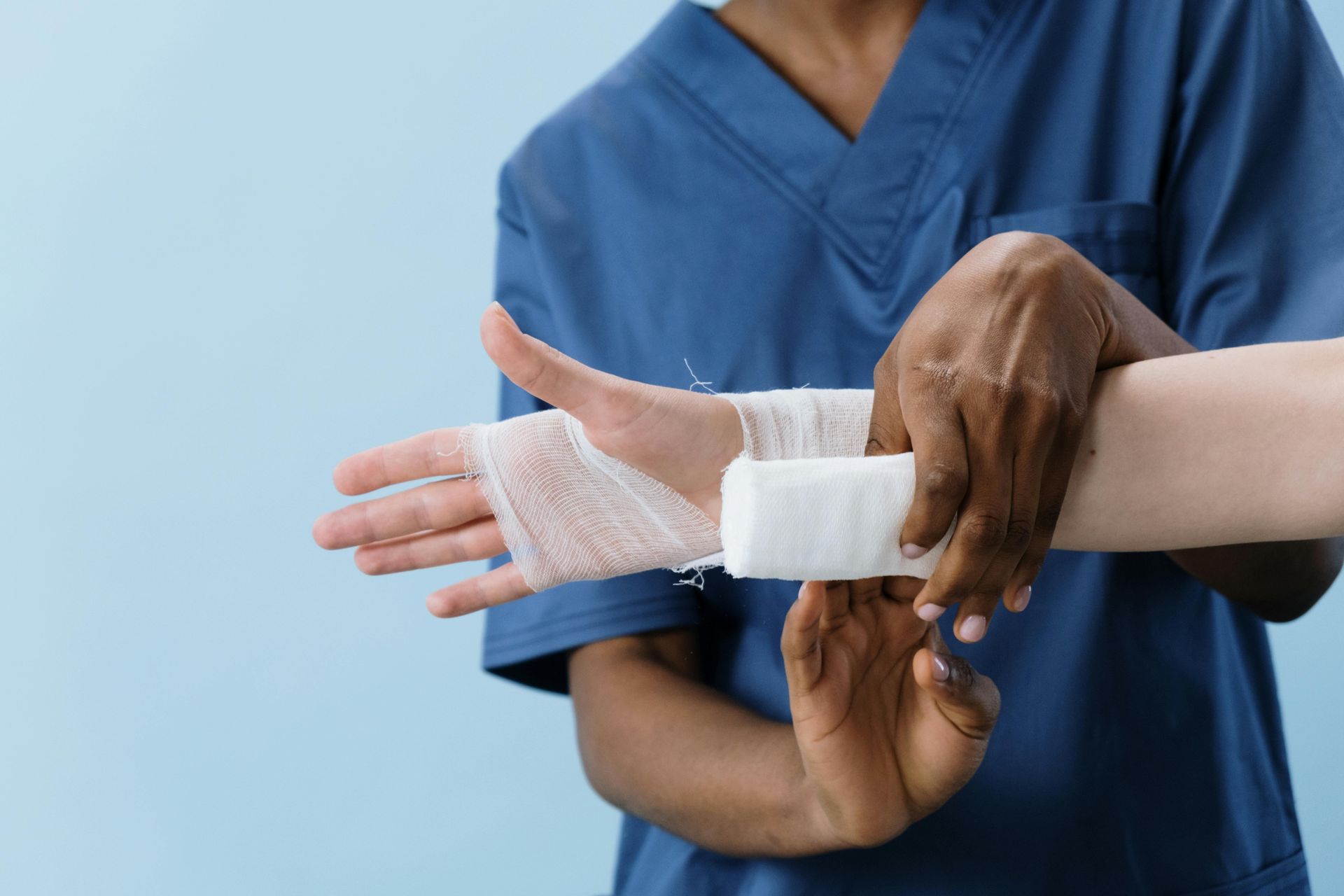 A person in blue scrubs applies a white medical bandage to another person's hand and wrist against a light blue background.