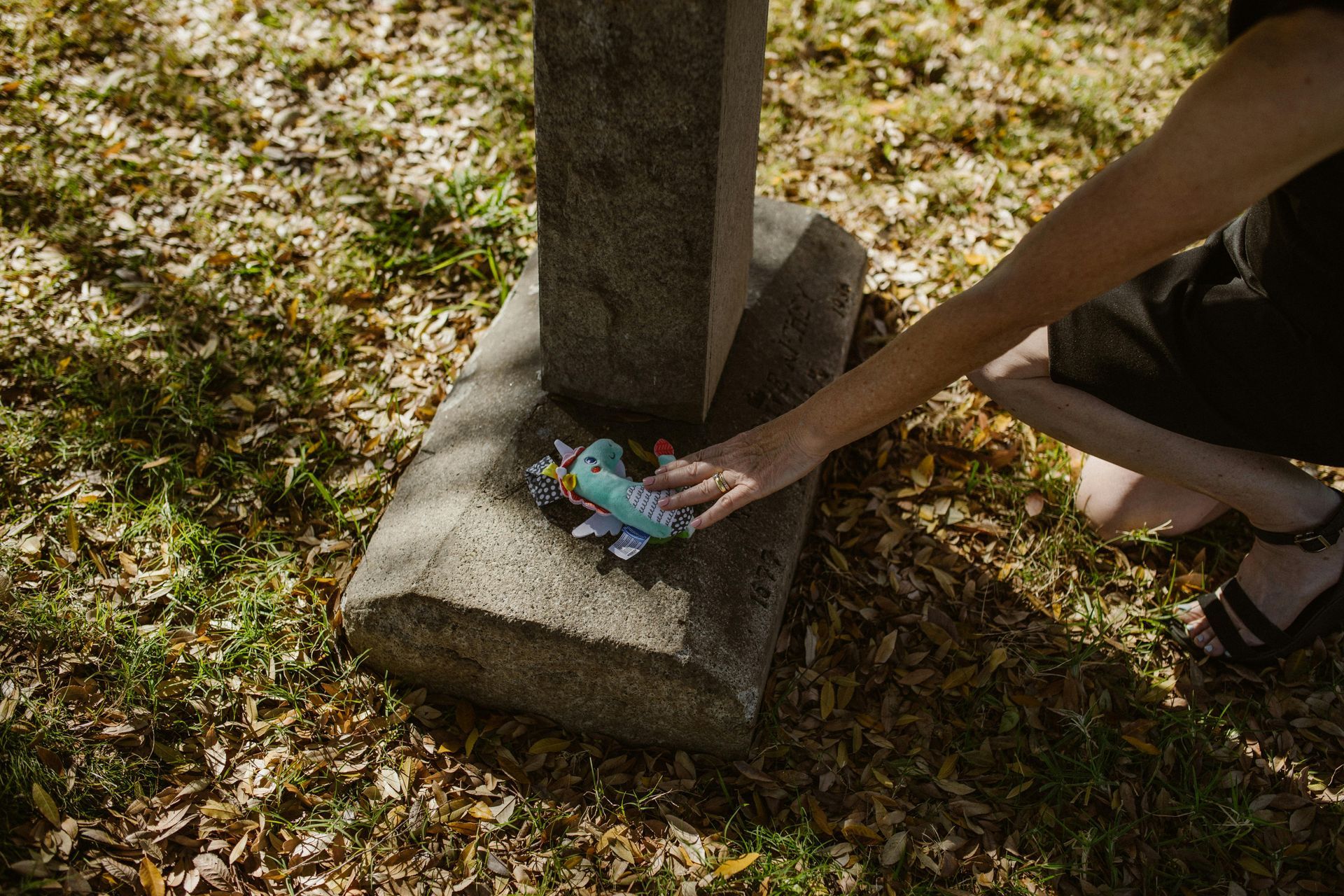 A person kneeling by a gravestone, placing a small, light-blue plush toy at its base on a leaf-covered lawn.