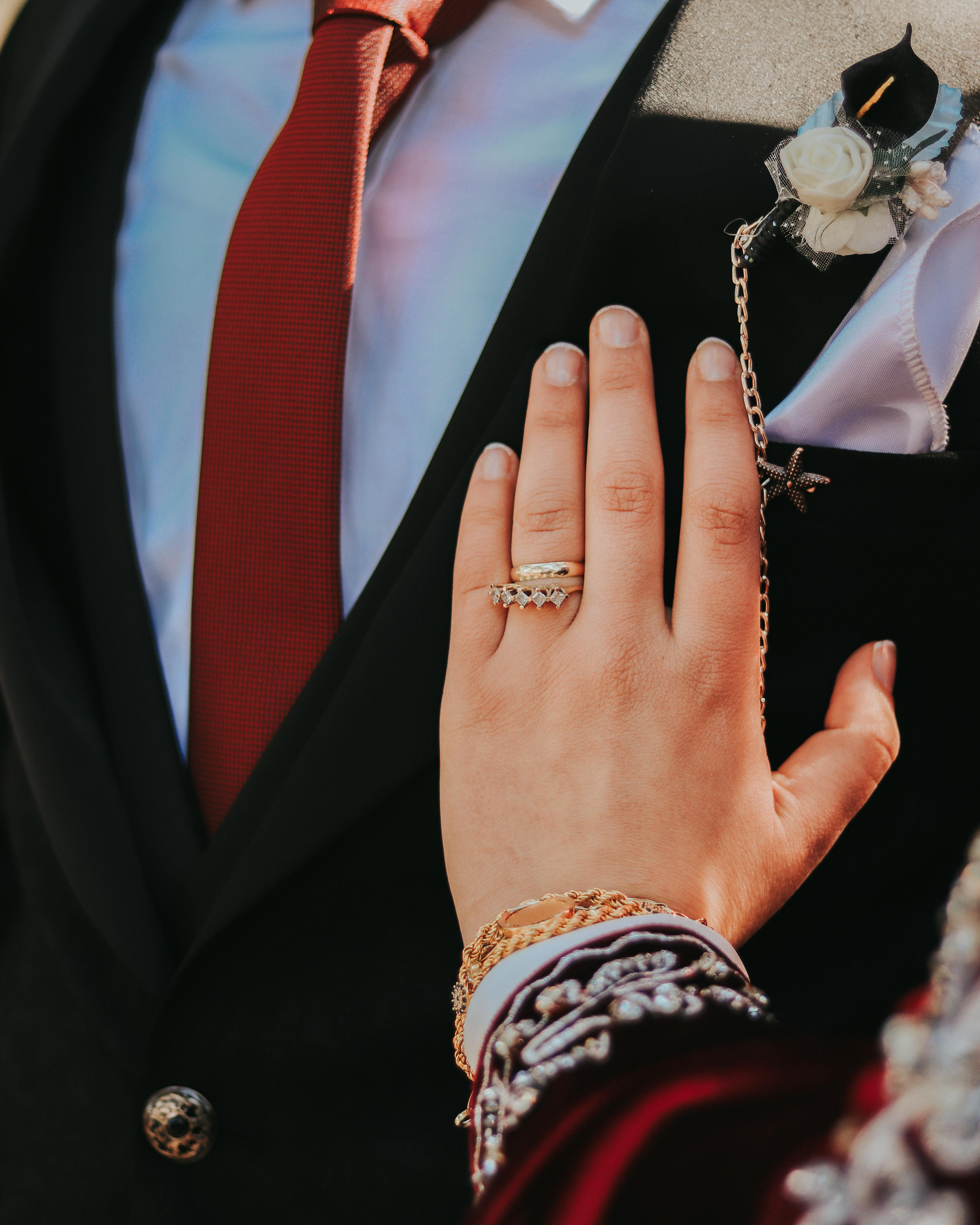 A hand with rings rests on the chest of a dark suit with a red tie and a small floral lapel pin.