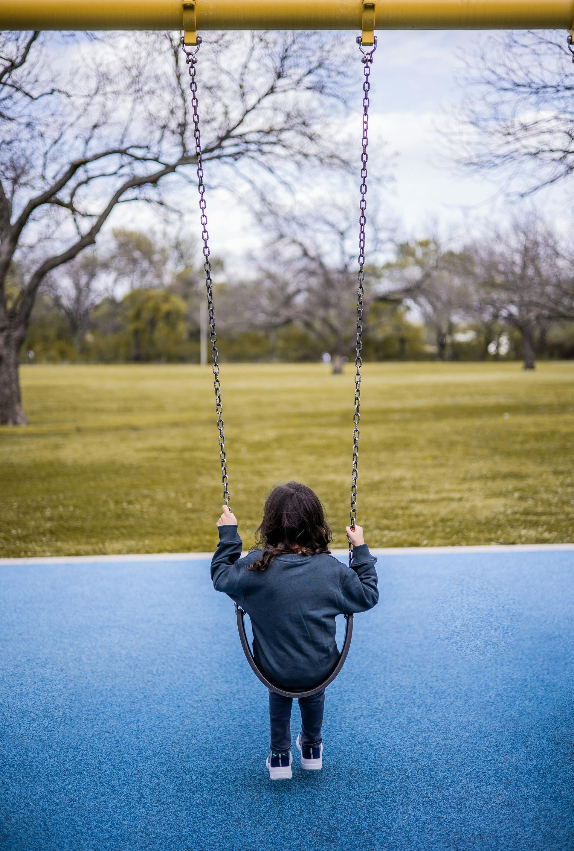 A person with long dark hair sits on a swing set facing away from the camera, over a blue surface with a field behind.