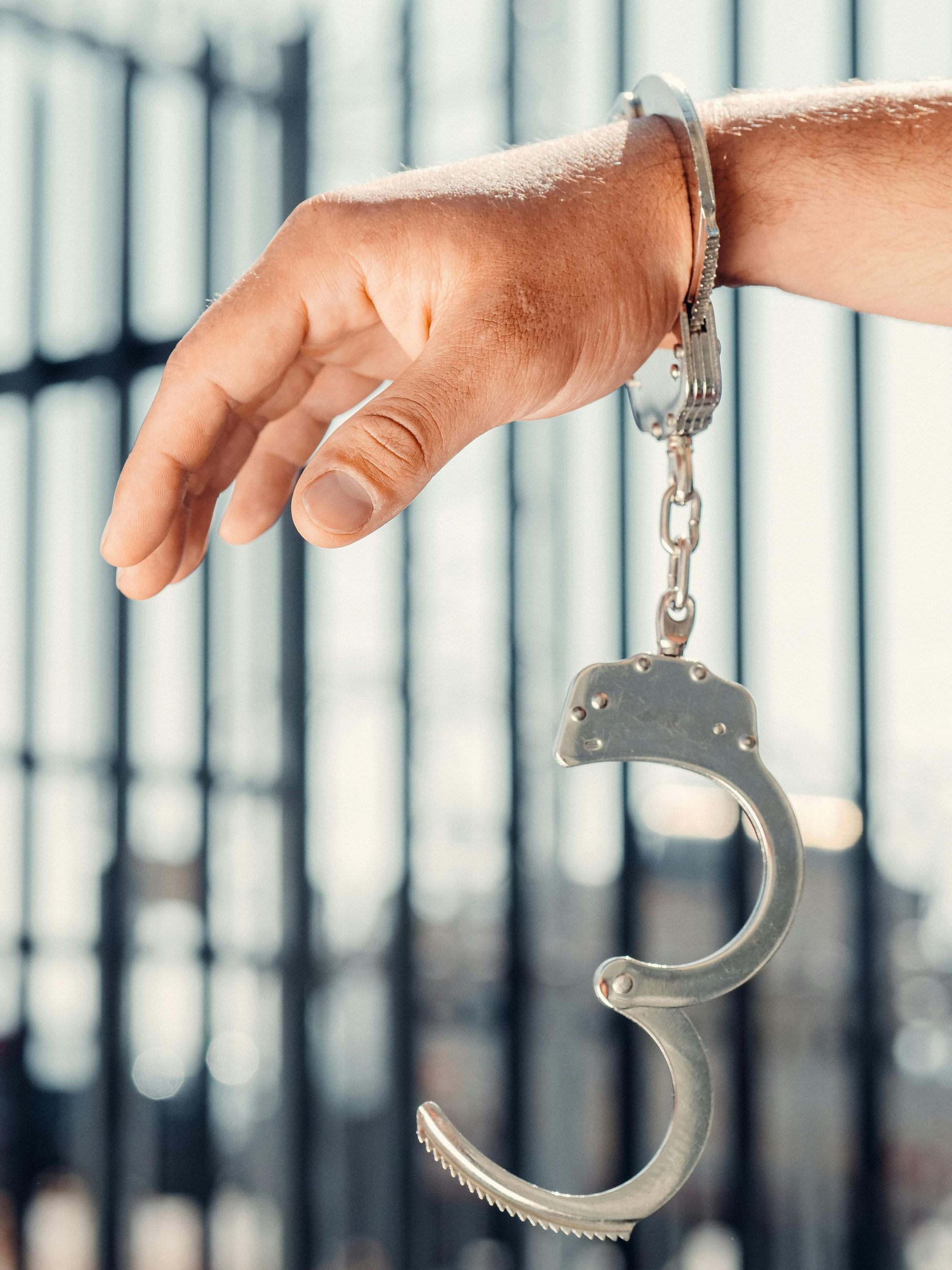 A hand hangs open with one handcuff locked on the wrist and the other hanging down, set against a blurry jail cell gate.