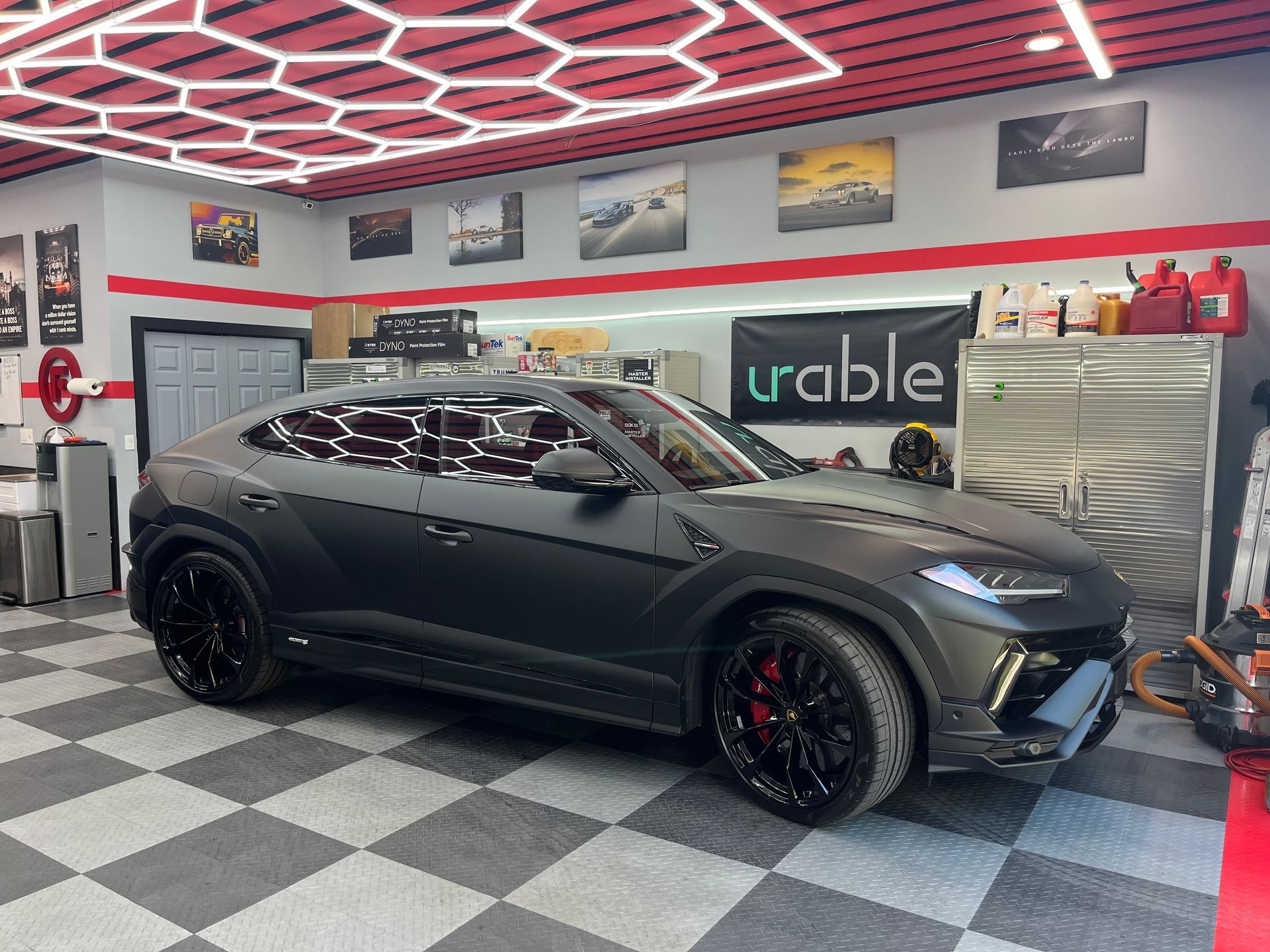 Matte black SUV in a custom garage with checkered floor, red hex ceiling lights, and car posters on the wall