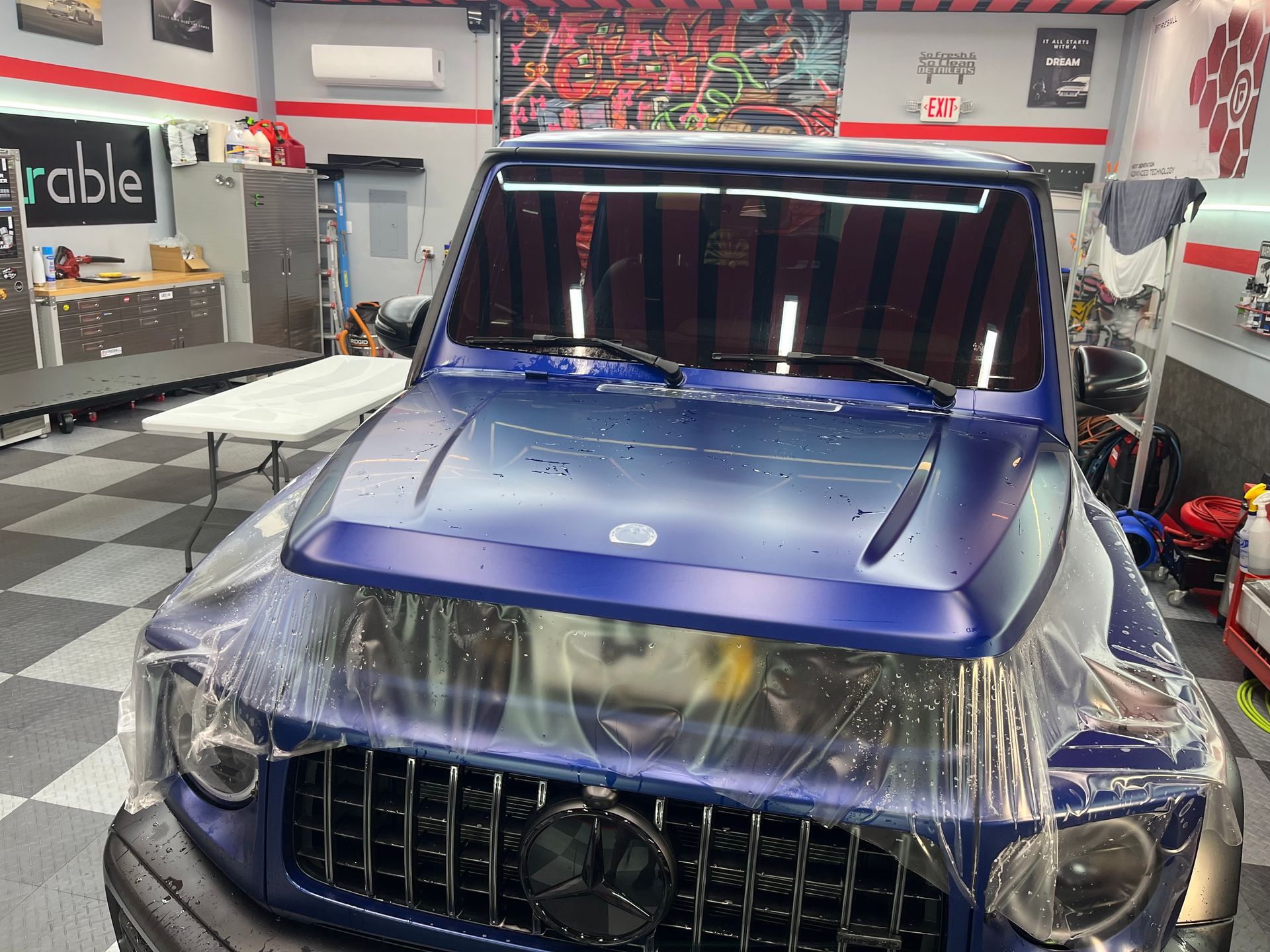 Blue SUV wrapped in protective plastic in a garage workshop, front view.