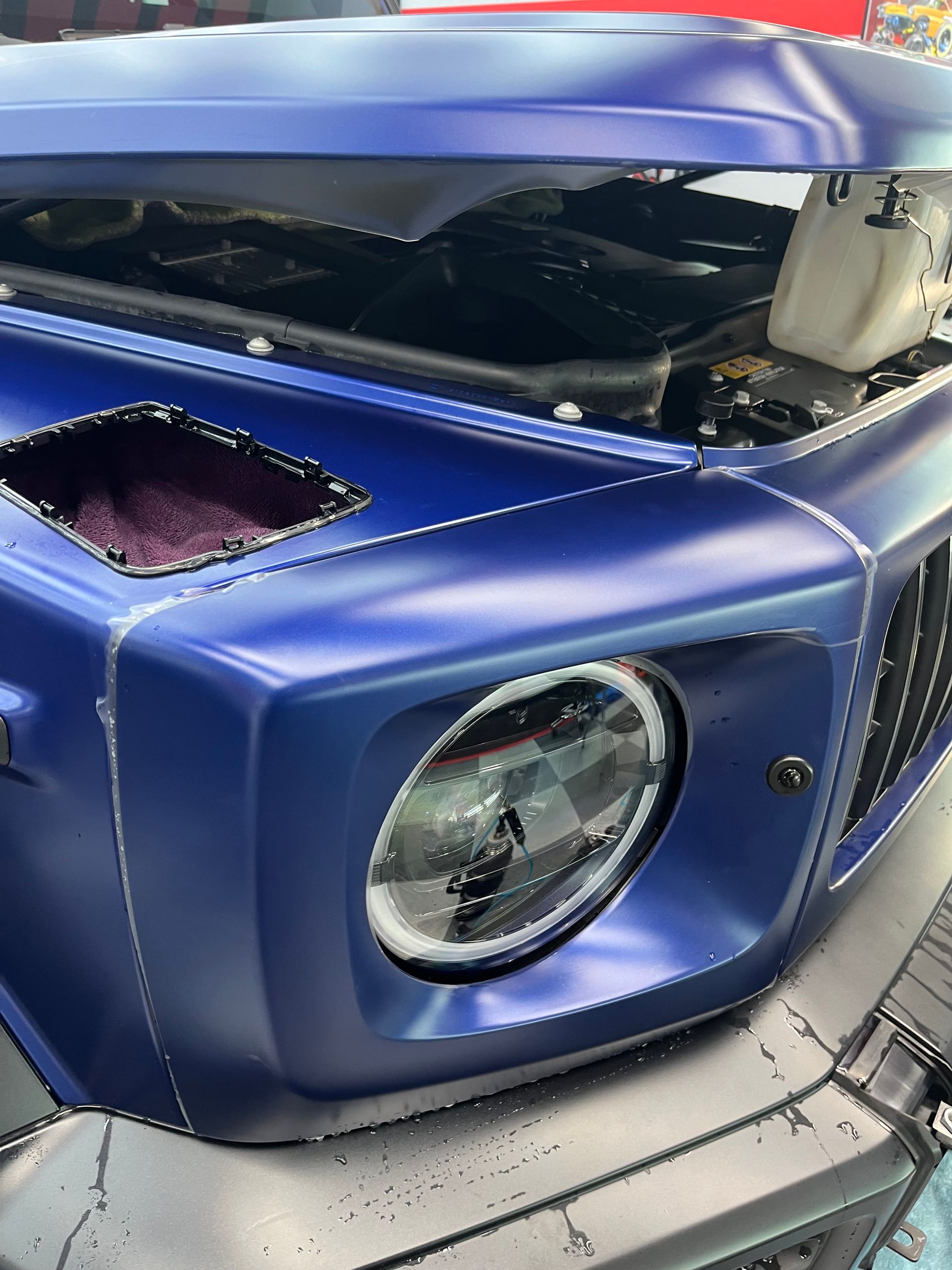 Blue vehicle front end with missing headlight and an open engine compartment