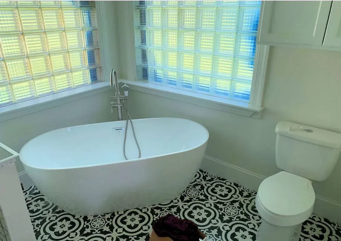 Bright bathroom with a freestanding tub, toilet, patterned floor, and sunlight through windows.