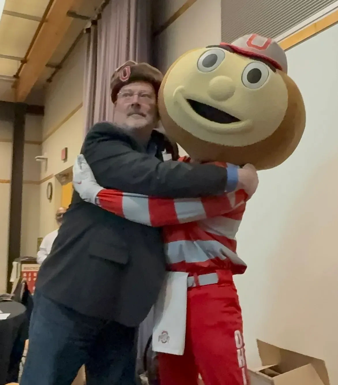 Man in a beret hugging a large smiling mascot in a red-and-white striped outfit indoors