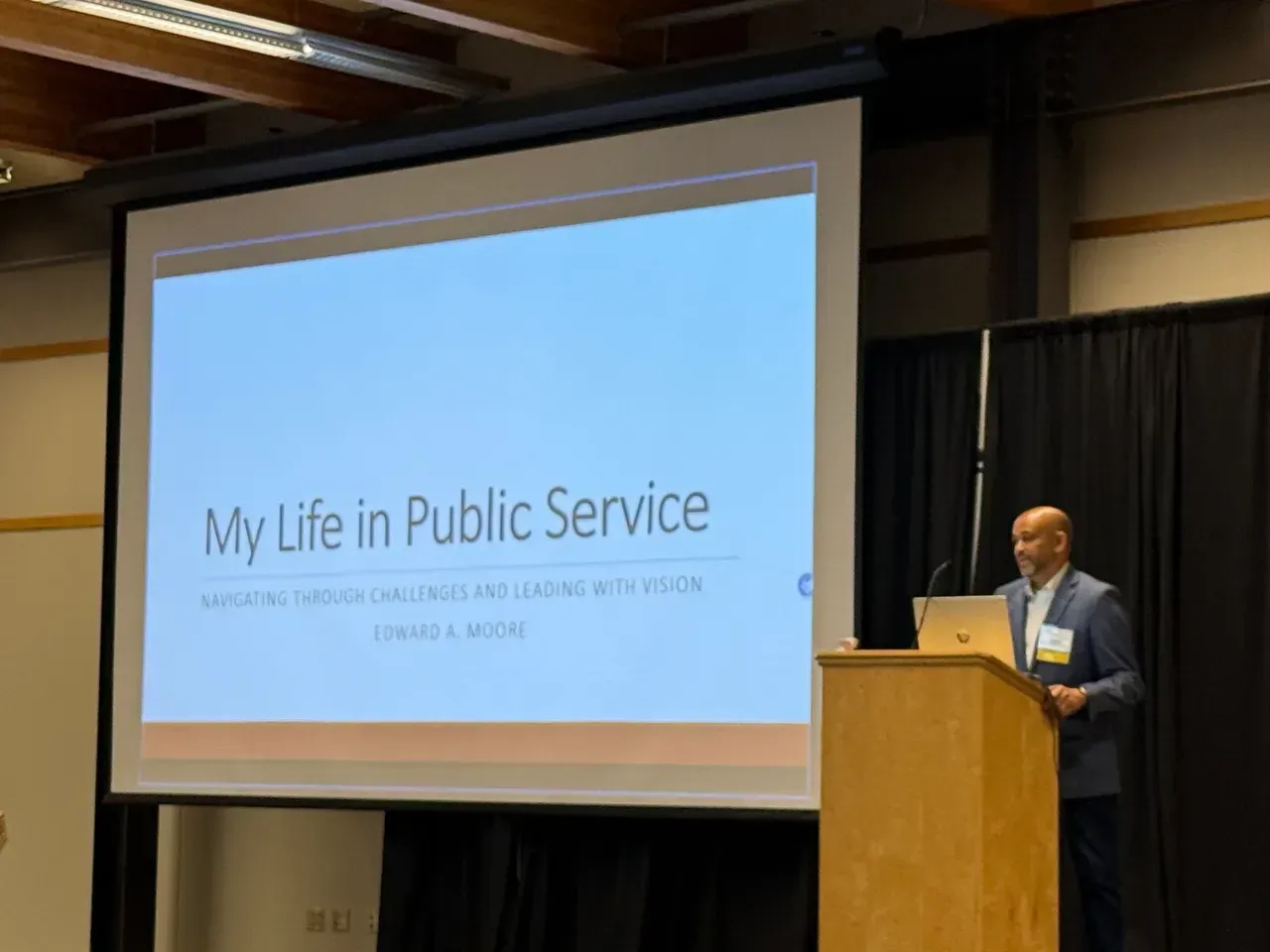 Speaker at podium beside slide titled “My Life in Public Service” in a conference room