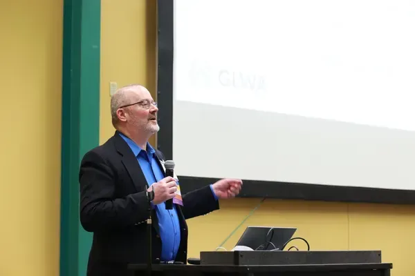 Man speaking at a podium during a presentation, gesturing toward a projection screen