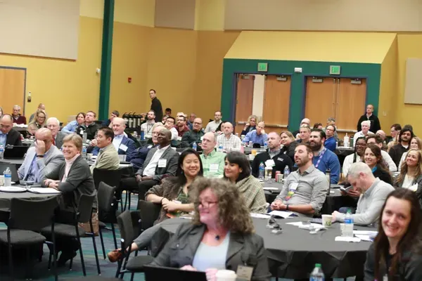Audience seated at round tables in a conference room, listening and smiling.