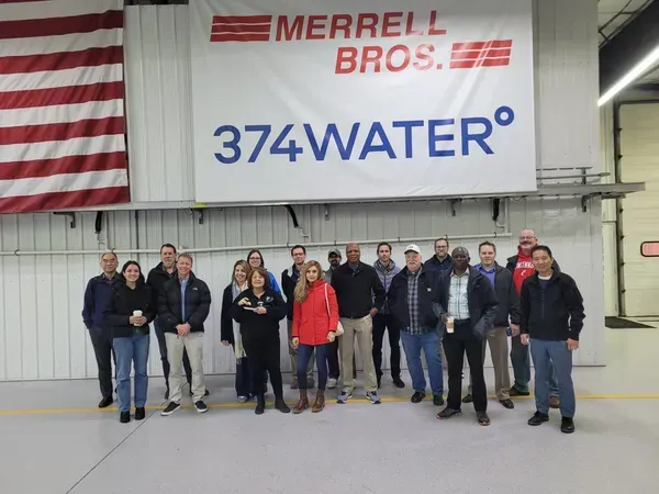 Group of people standing indoors under a Merrell Bros. 374WATER banner and an American flag.