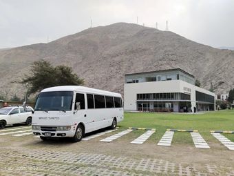 Minibús blanco frente a un edificio moderno, con una colina rocosa al fondo y una zona de aparcamiento con césped.