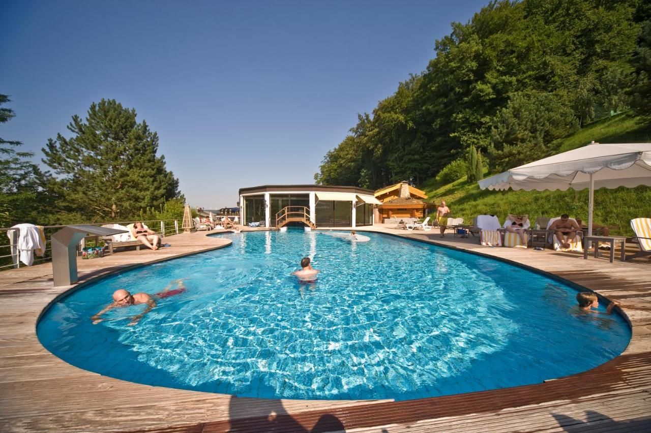 Une piscine extérieure ensoleillée où les gens prennent un bain de soleil. Elle est entourée d'une végétation luxuriante.
