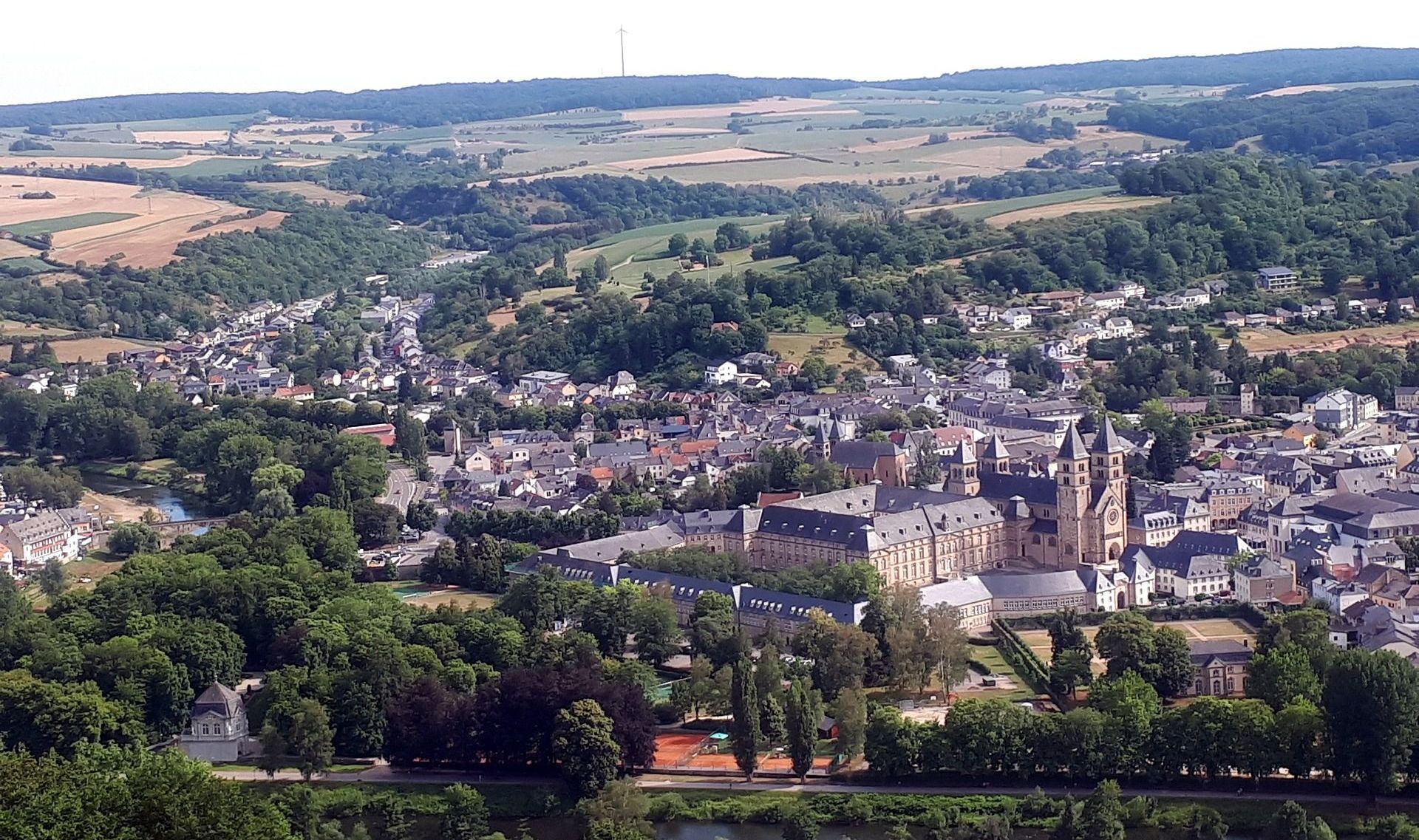 Vue aérienne d'une petite ville européenne avec des bâtiments en pierre, entourée d'arbres et de collines sous un ciel bleu.