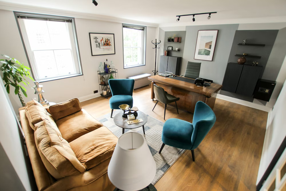 Cozy office space with a brown couch, two teal chairs, a large wooden desk, and two windows.