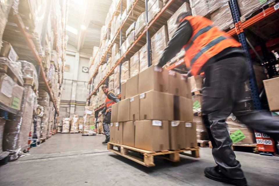 I magazzinieri con i giubbotti arancioni spostano scatole di cartone su un pallet attraverso un magazzino affollato.