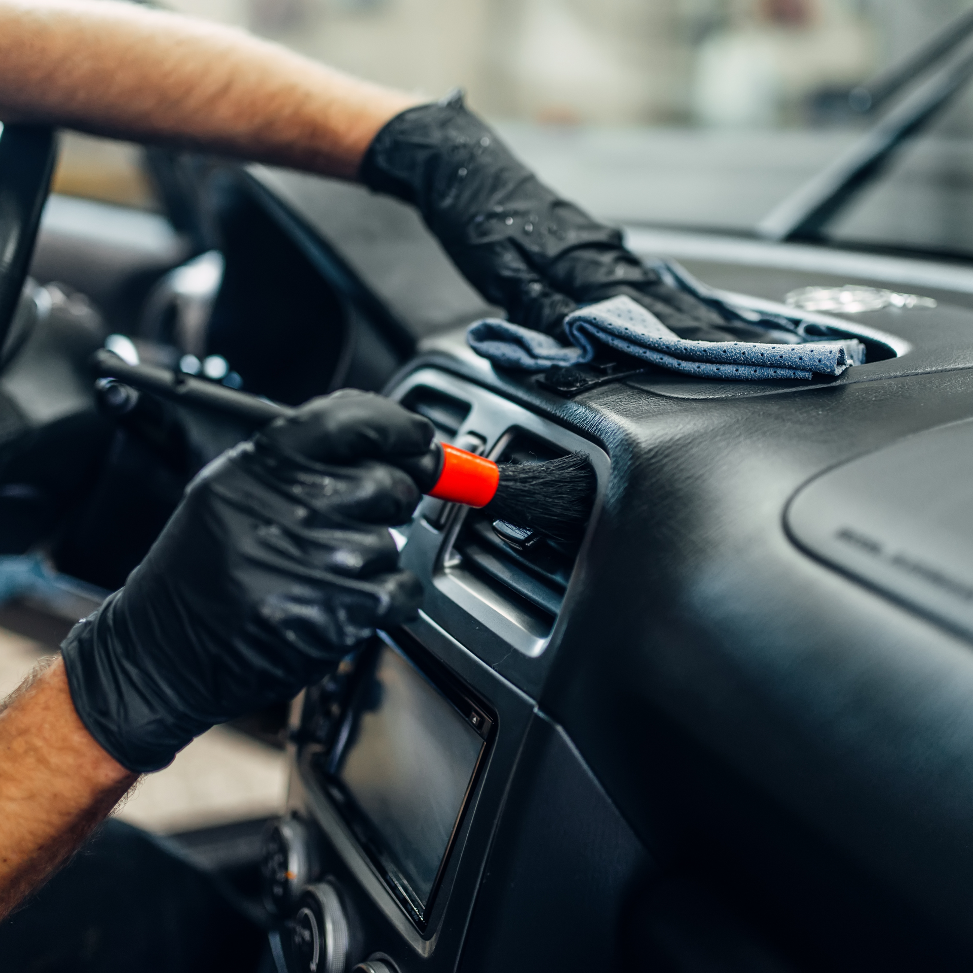 Person cleaning a car dashboard with a brush and cloth, wearing black gloves.