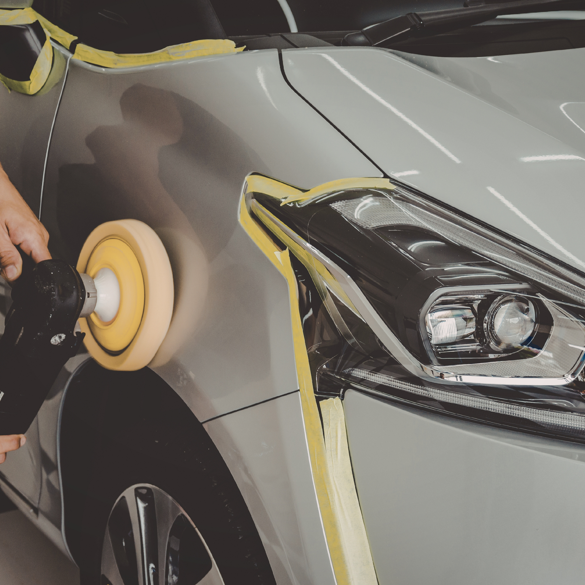 Person polishing a silver car with a buffing machine, yellow tape protecting edges.