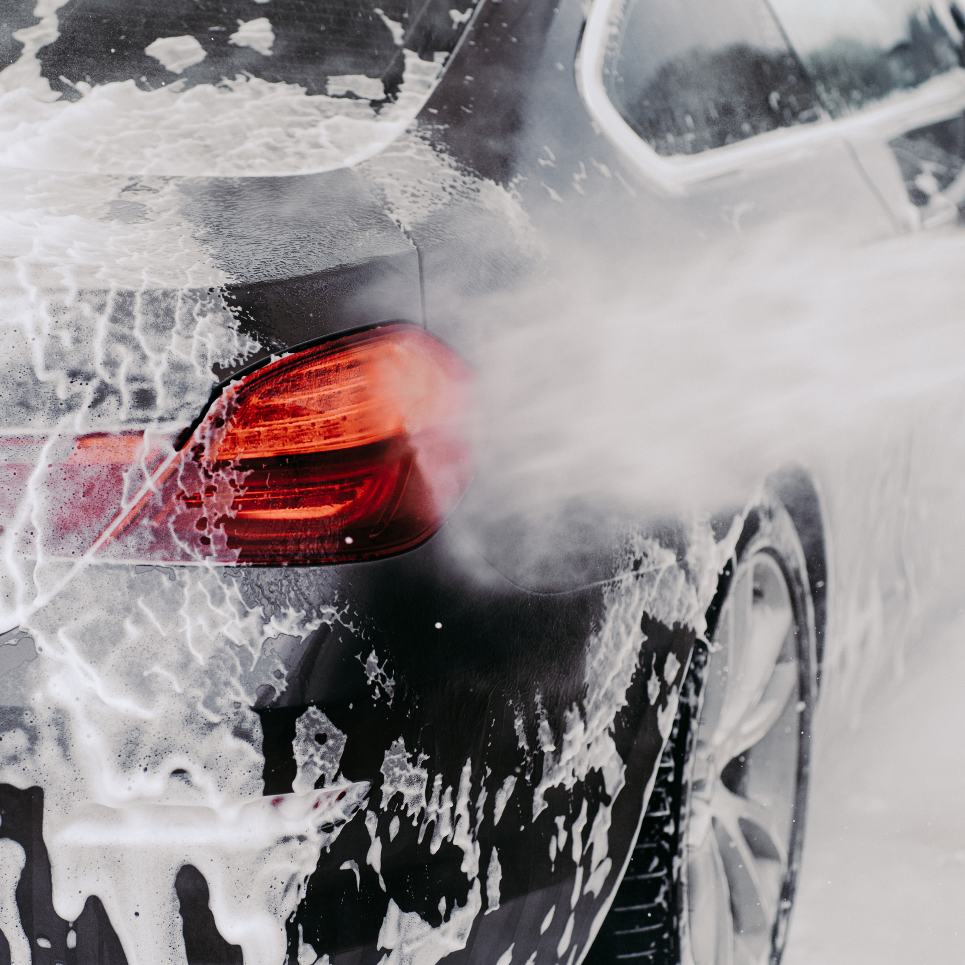Car being washed with foamy soap. Spraying water near tail light and tire.