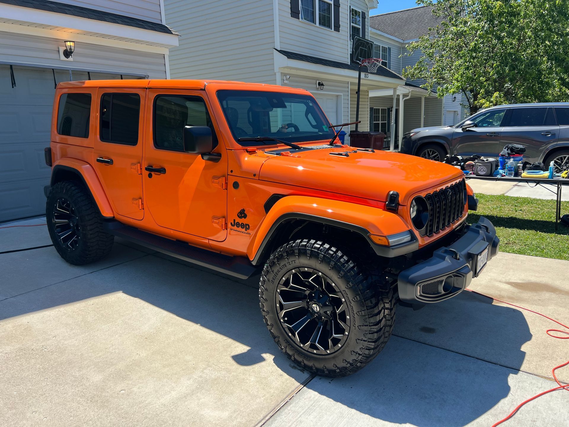 Orange Jeep Wrangler parked in a driveway outside a house, with black wheels and lifted tires