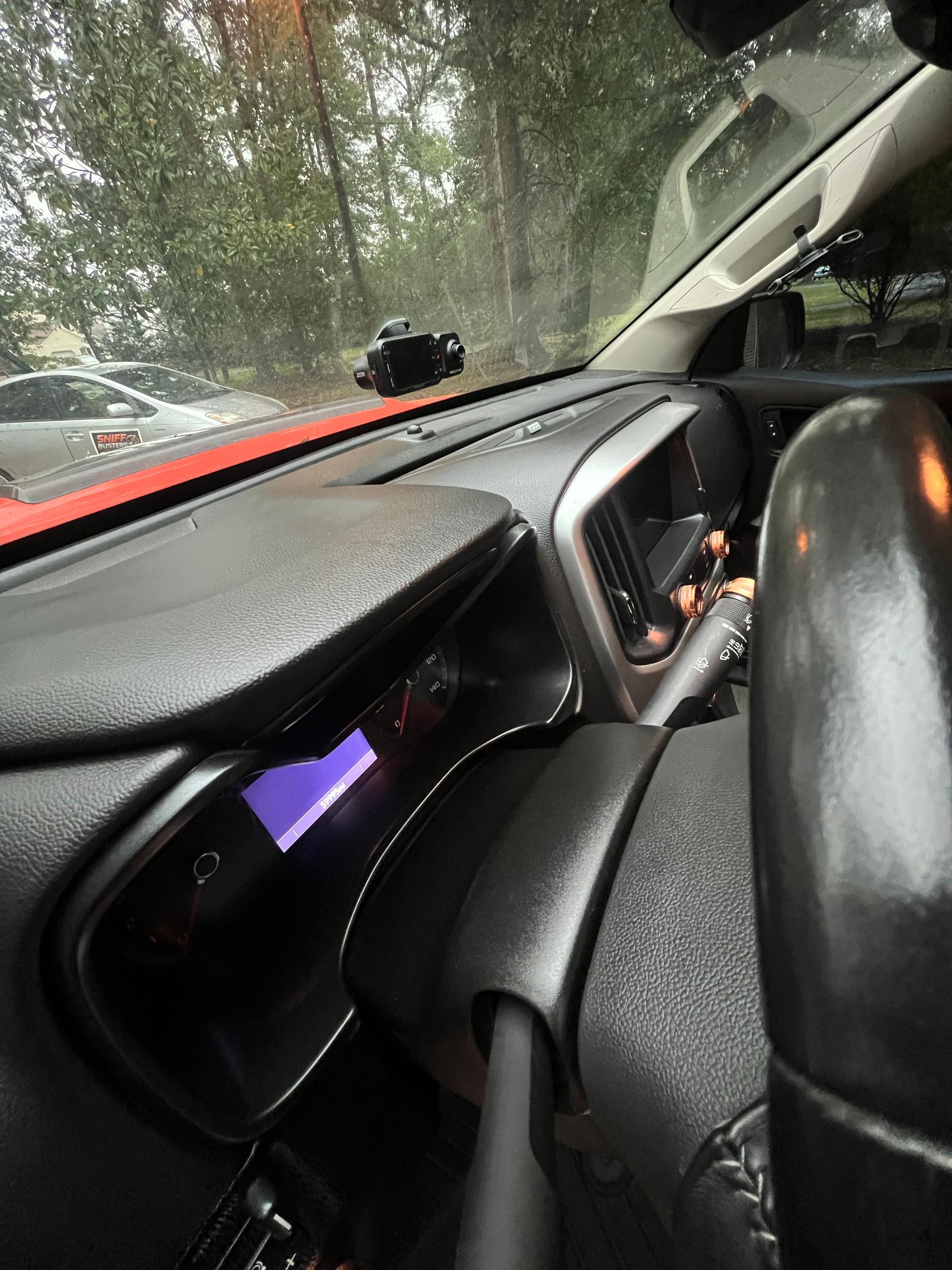 Car interior with dashboard and steering wheel, rain-speckled windshield, and a small purple-lit display.